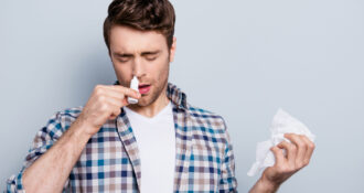 Portret atrakcyjnego mężczyzny z aerozolem do nosa i chusteczką w rękach, używającego kropli do nosa na szarym tle, koncepcja leczenia alergii lub przeziębienia, styl zimowy.