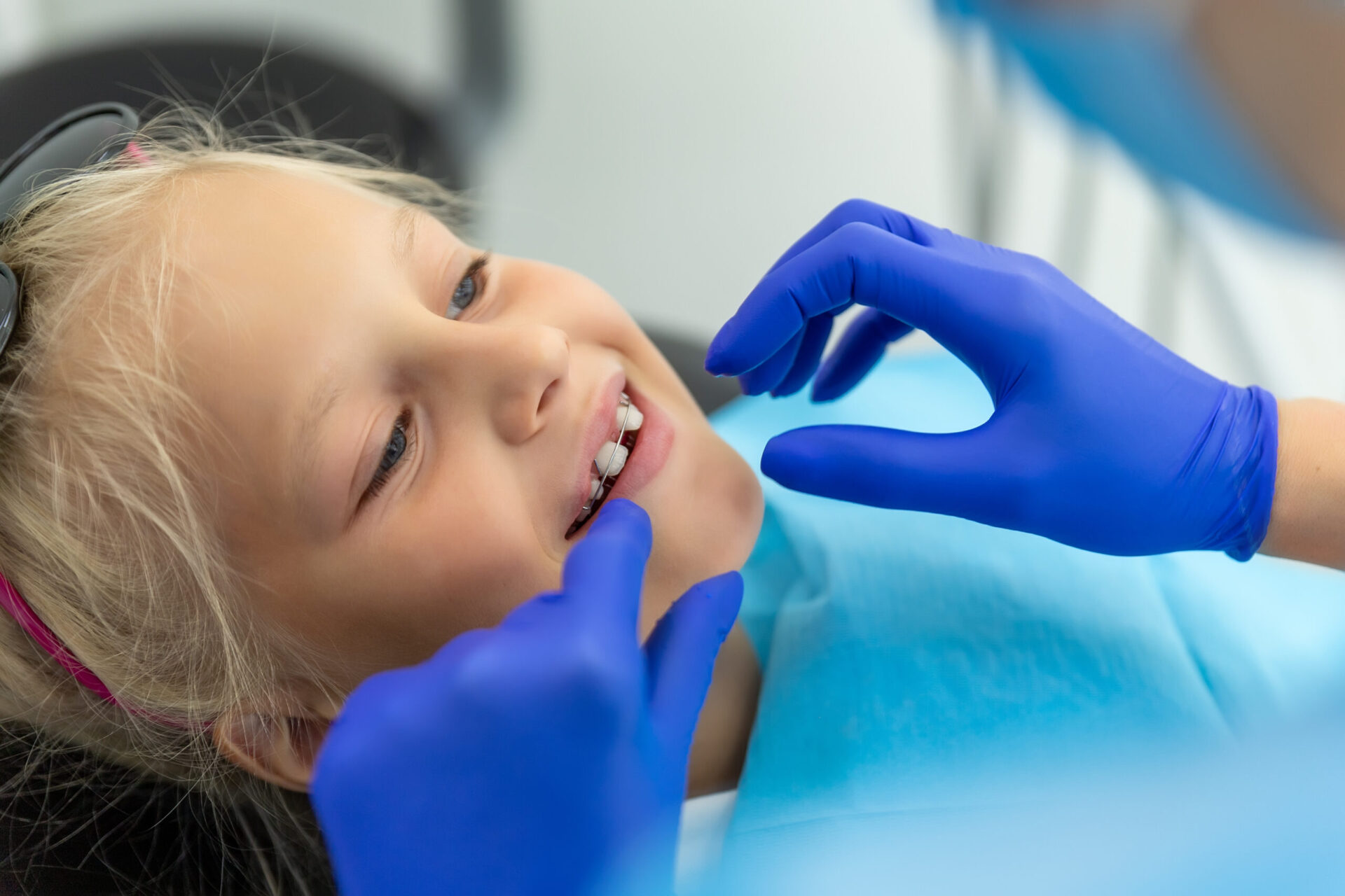 Mała urocza, szczęśliwa, blondynka dziewczynka pochodzenia kaukaskiego u dentysty podczas wizyty kontrolnej związanej z aparatami ortodontycznymi. Dziecko podczas wizyty u ortodonty i leczenia jamy ustnej. Opieka i higiena jamy ustnej dzieci.