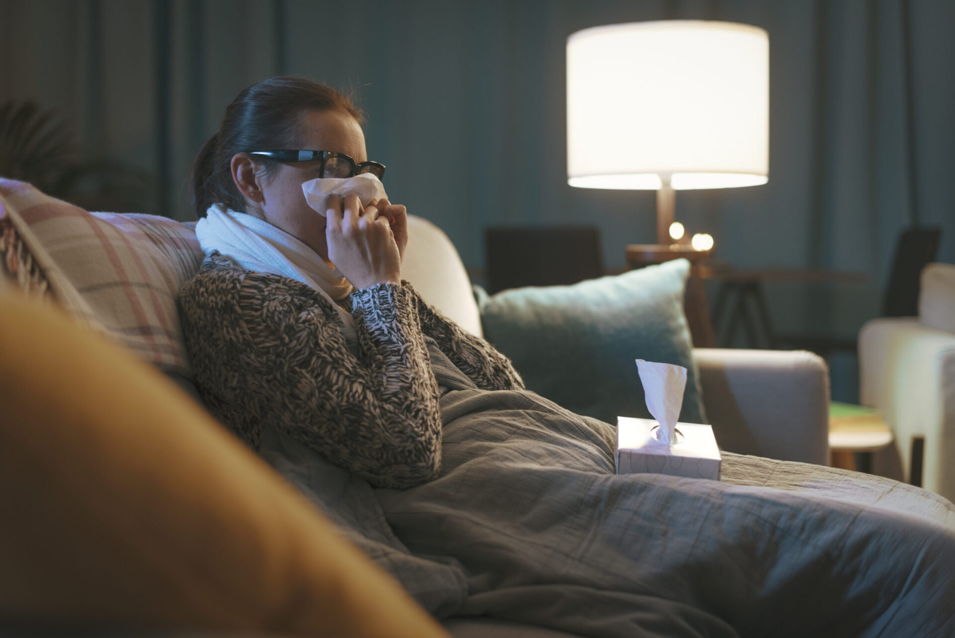 Chora kobieta z przeziębieniem i grypą odpoczywająca w domu na kanapie, dmuchająca nos.