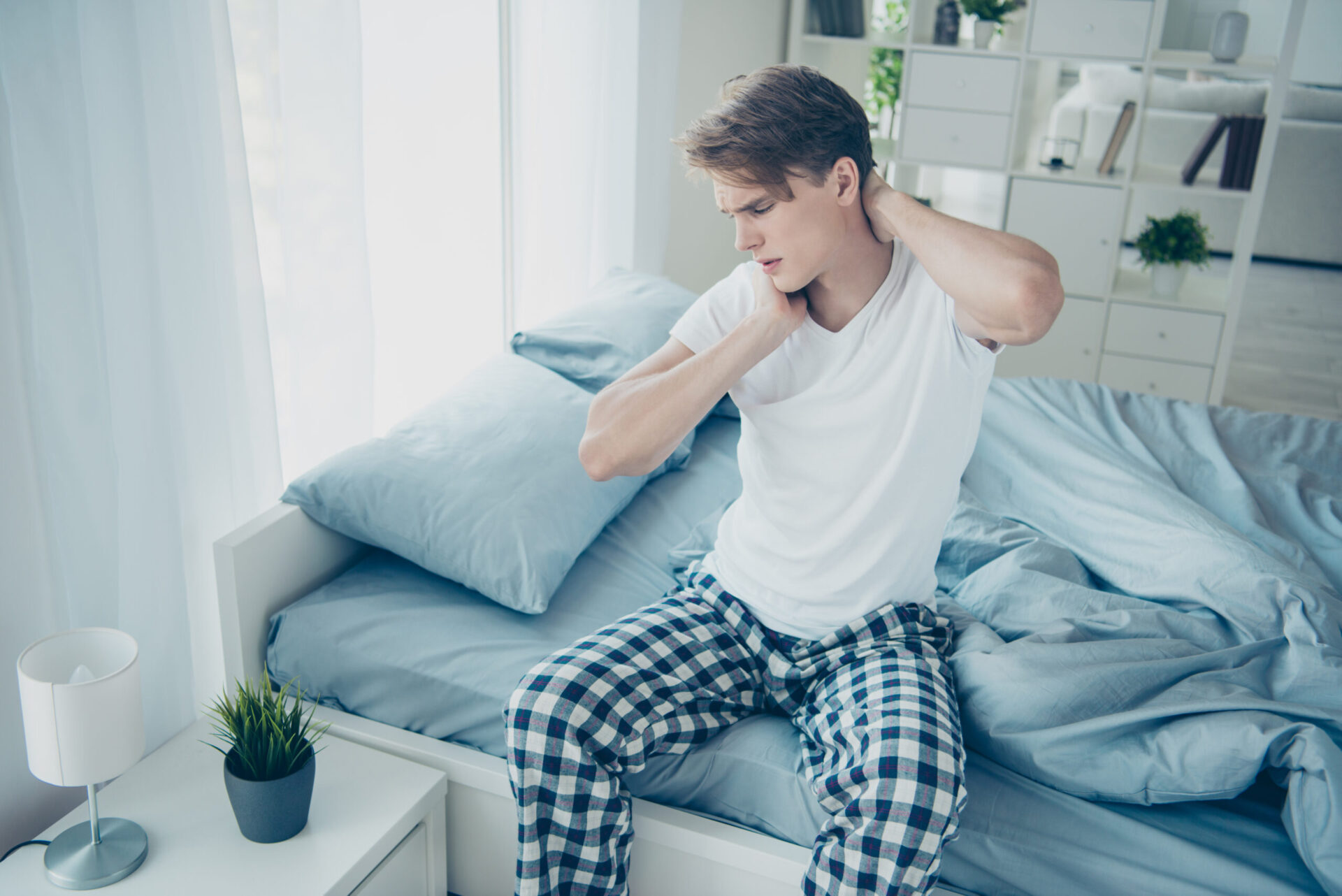 Portret zaniepokojonego, smutnego mężczyzny siedzącego na łóżku, obudzonego z bólem szyi i napięciem mięśni, ubranego w kraciaste białe piżamowe spodnie, w domu.