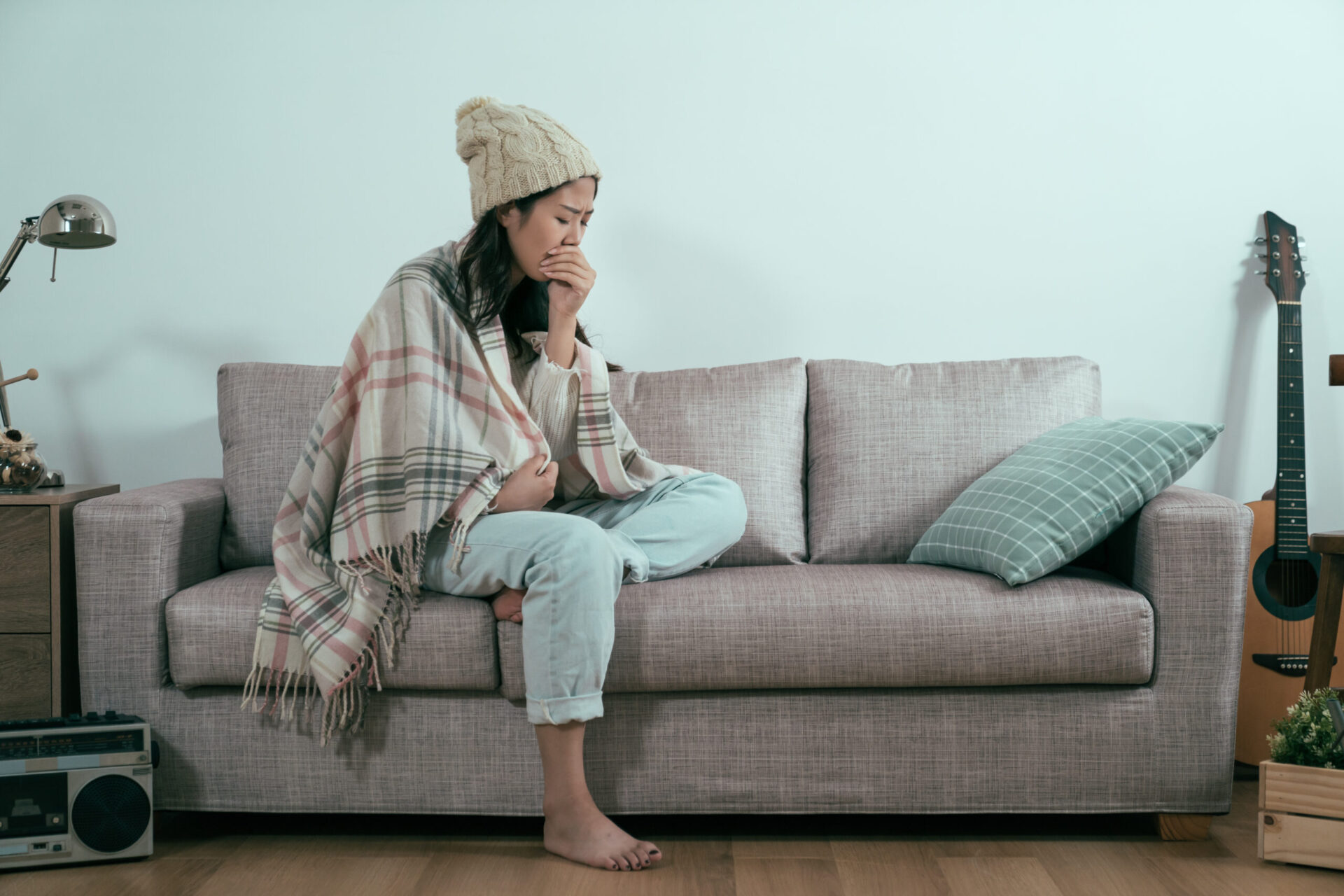 Kobieta o azjatyckim pochodzeniu w ciepłym swetrze, chora i kaszląca na białym tle. Chora dziewczyna w zimowej czapce z pomponem cierpiąca na przeziębienie na jasnej kanapie w salonie mieszkania.